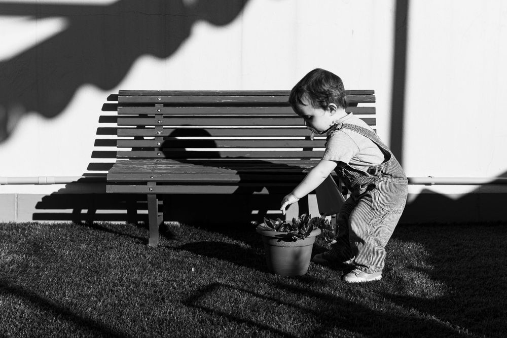 A small child in overalls plays near a wooden bench casting dramatic shadows in black and white.