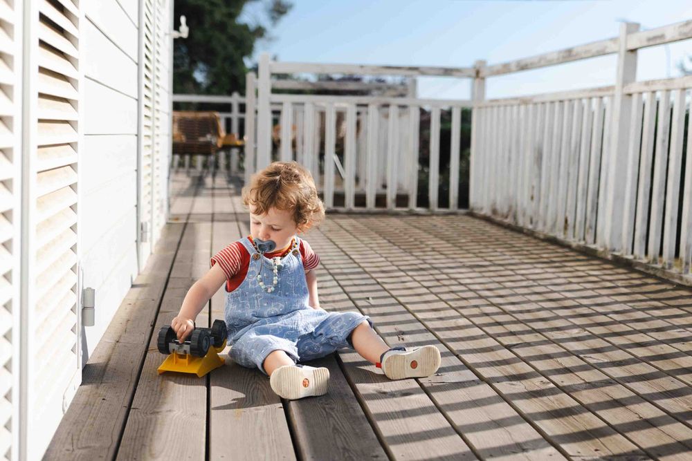 Small child in overalls sits playing with a toy truck on a wooden deck with white railings.