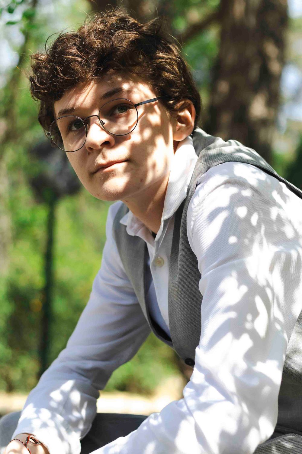 Person in glasses and white button-down shirt smiling while sitting outdoors in dappled sunlight.