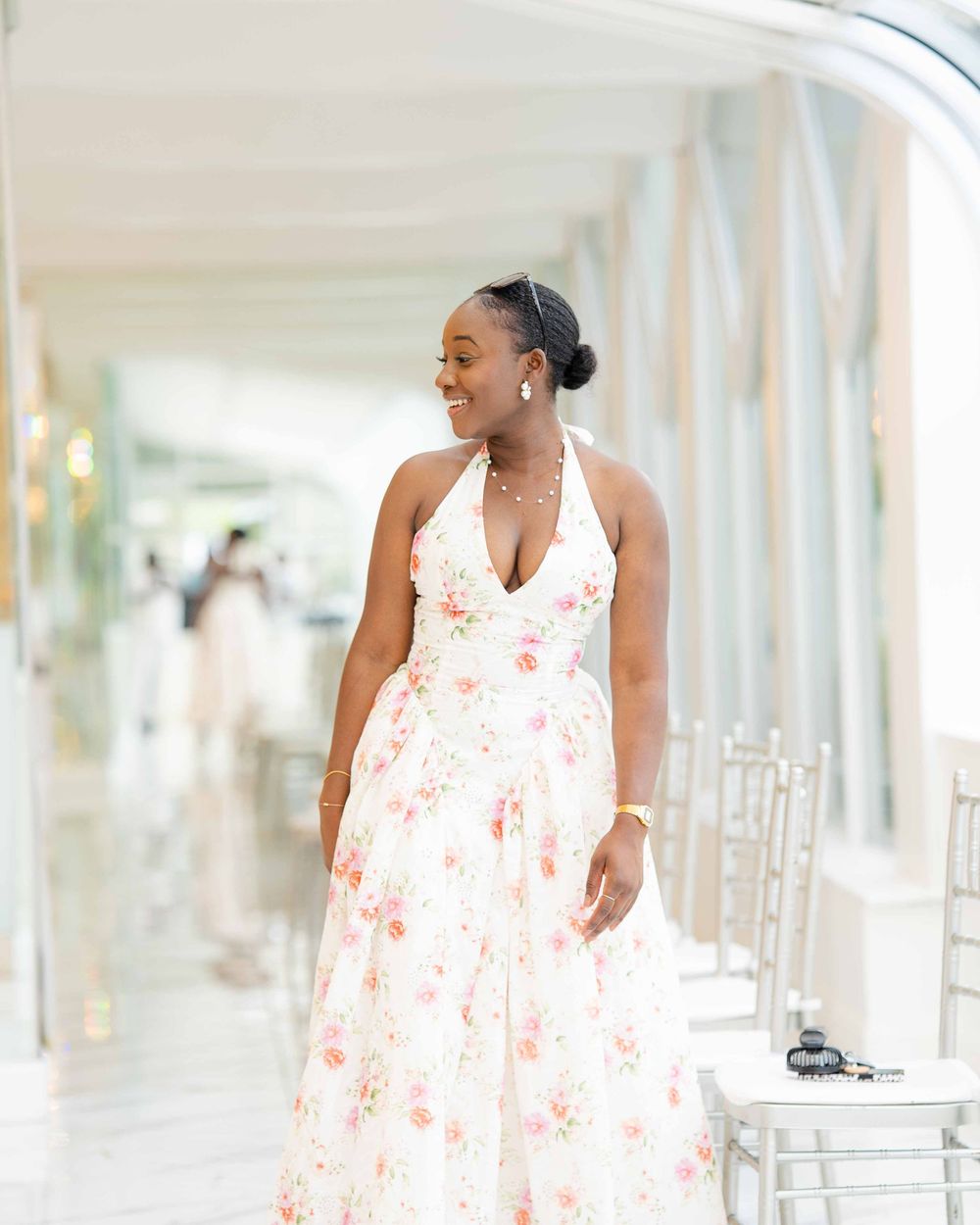 Someone in a flowing white halter dress with delicate floral print poses elegantly in a bright hallway setting.