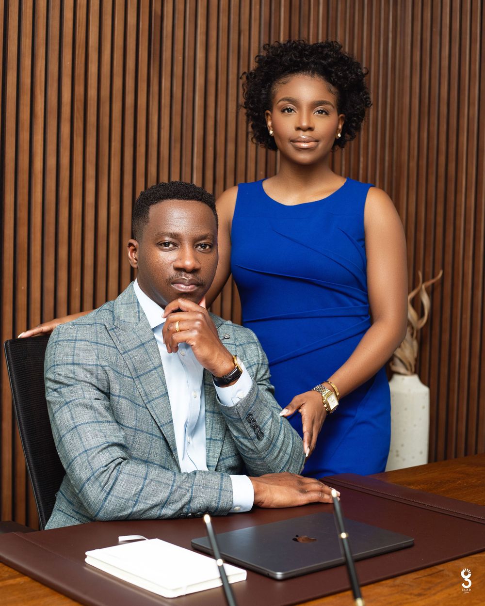Professional portrait of two business colleagues in formal attire posing against a wood-paneled wall in an office setting.