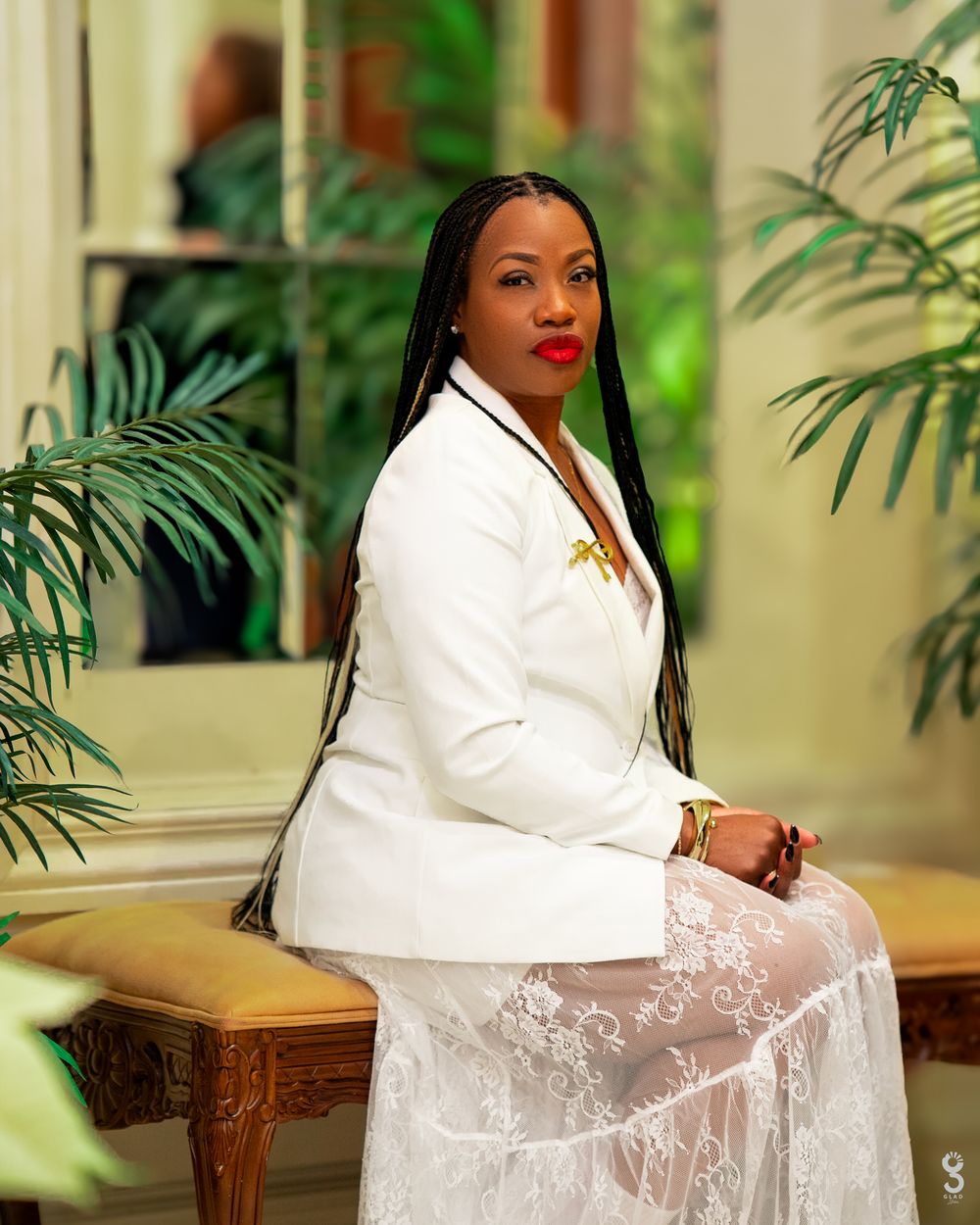 Professional portrait in elegant white blazer and lace skirt seated on ornate bench with tropical plants in background.