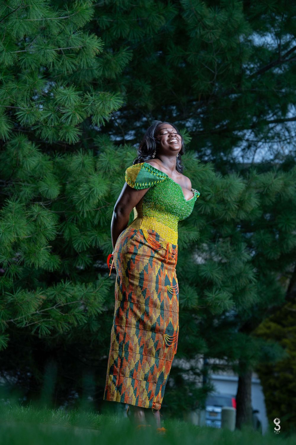 A person wearing a vibrant yellow and orange patterned dress stands against a backdrop of lush green pine trees.