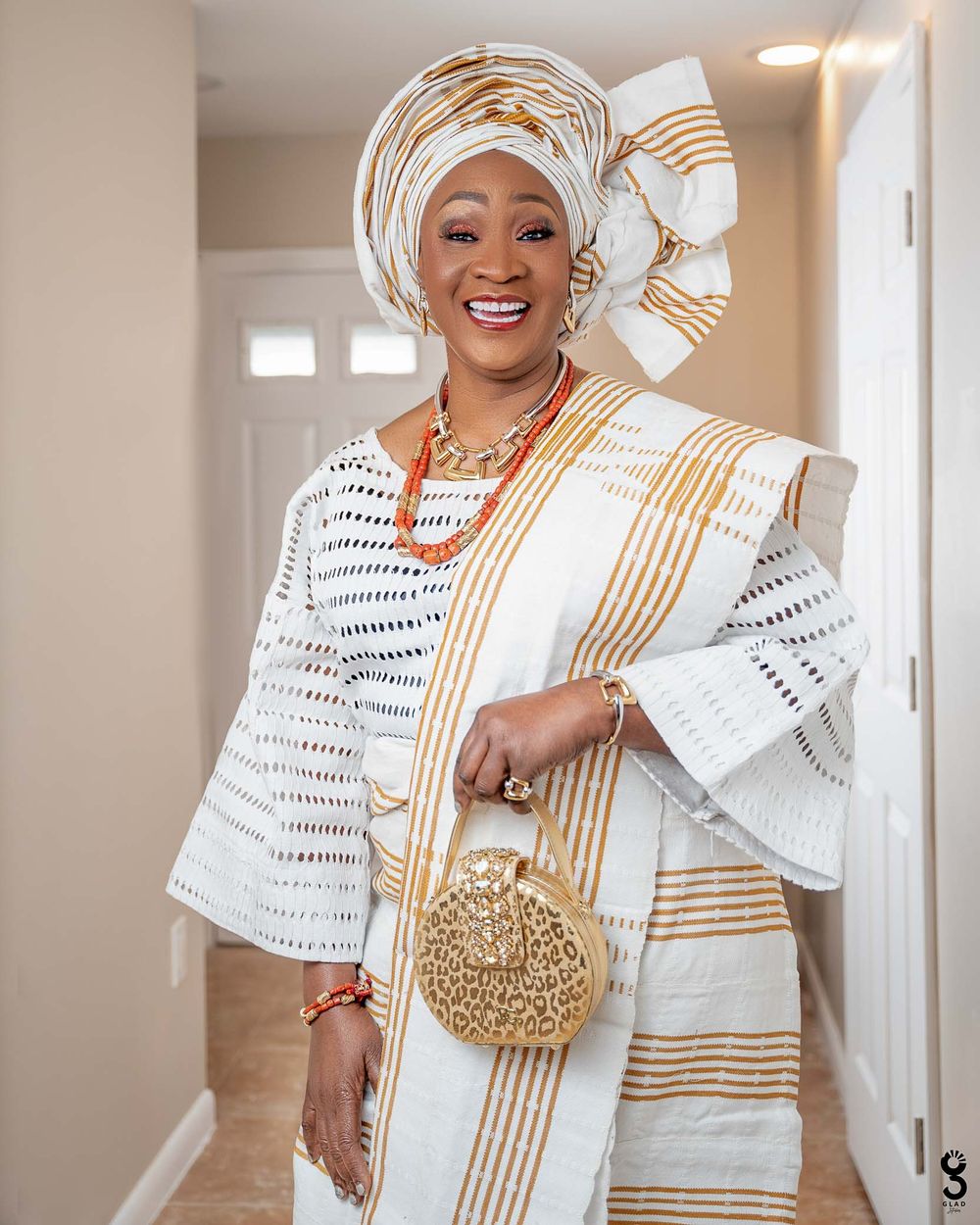 Woman in elegant white and gold African traditional attire with headwrap poses joyfully in hallway.