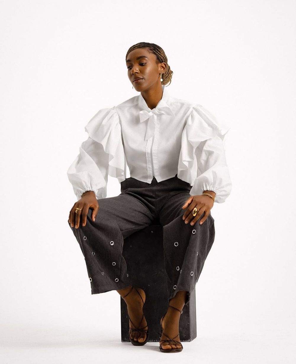 Fashion model in white puff sleeve blouse and black pants poses on cube against white backdrop.