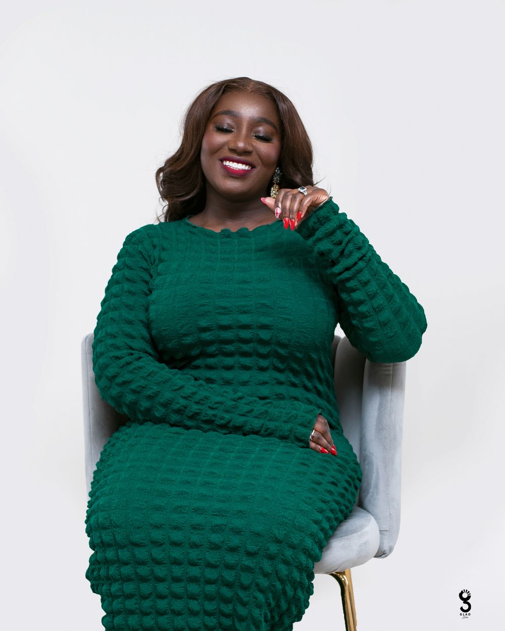 Someone wearing a textured emerald green sweater dress sits smiling in a light gray chair against a white background.