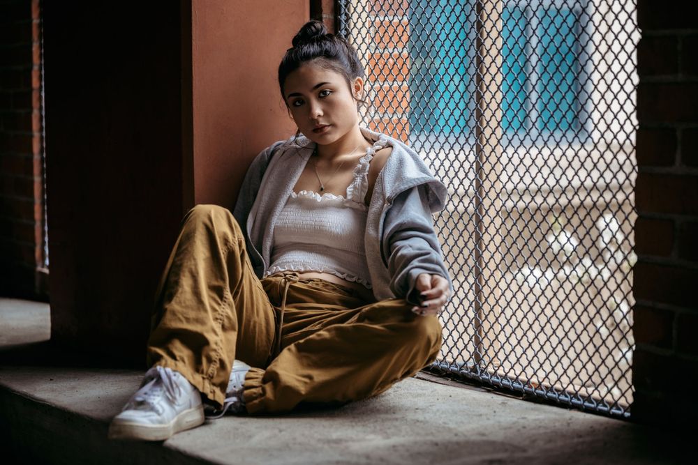 Person in casual streetwear sits against wall by chain-link fence in moody portrait with blue and orange tones.