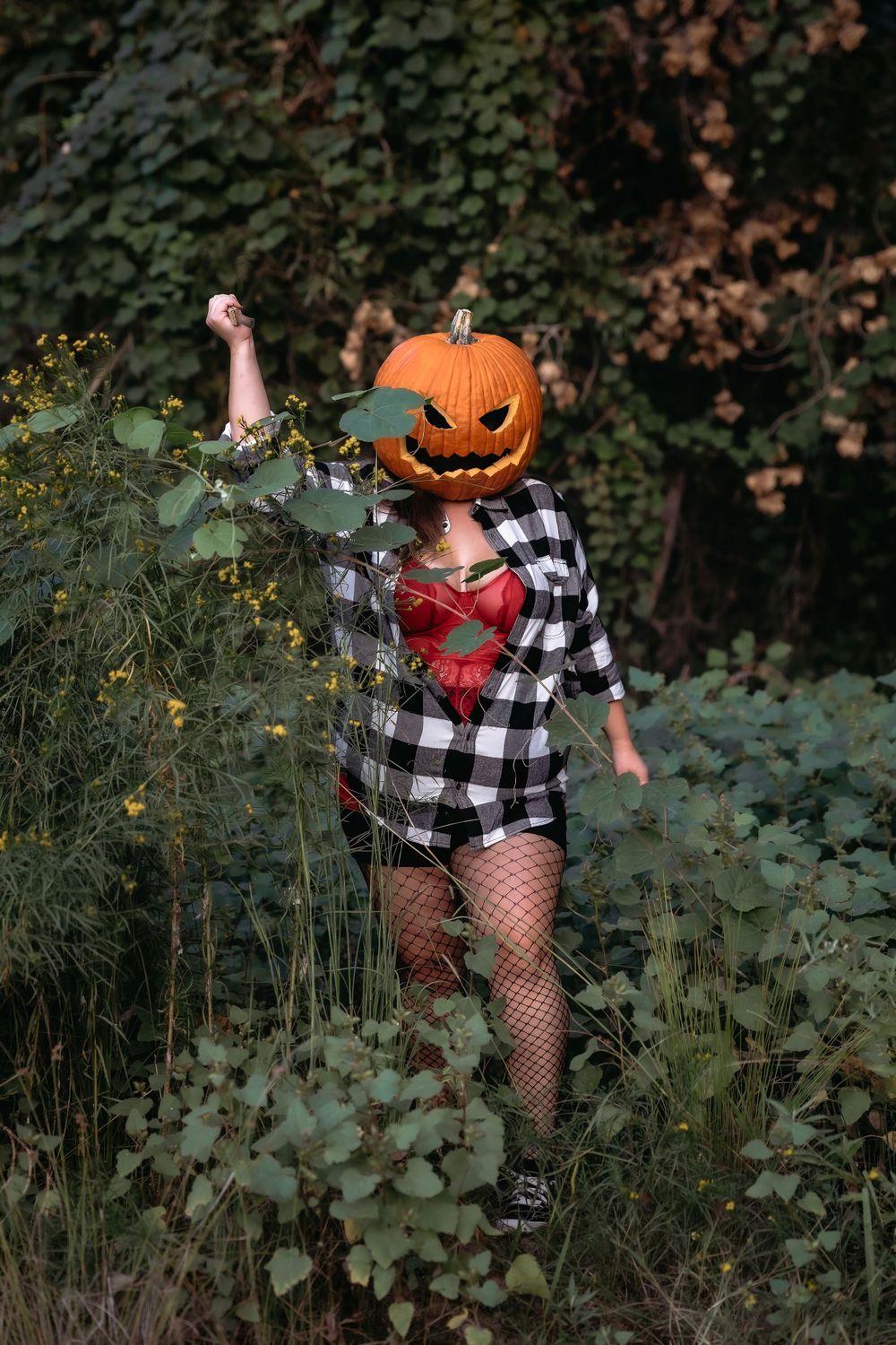 A pumpkin-headed figure in a plaid shirt and fishnet stockings poses dramatically in a garden setting during autumn.