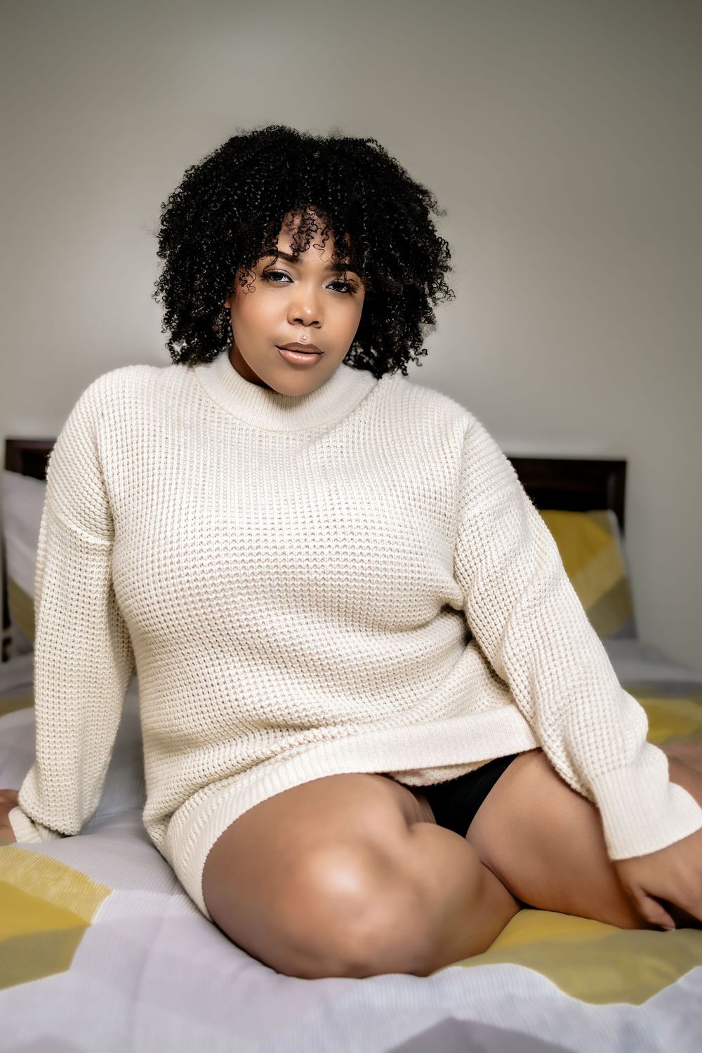 A person wearing a cozy cream-colored sweater relaxes on bedding with yellow accents and natural curly hair.