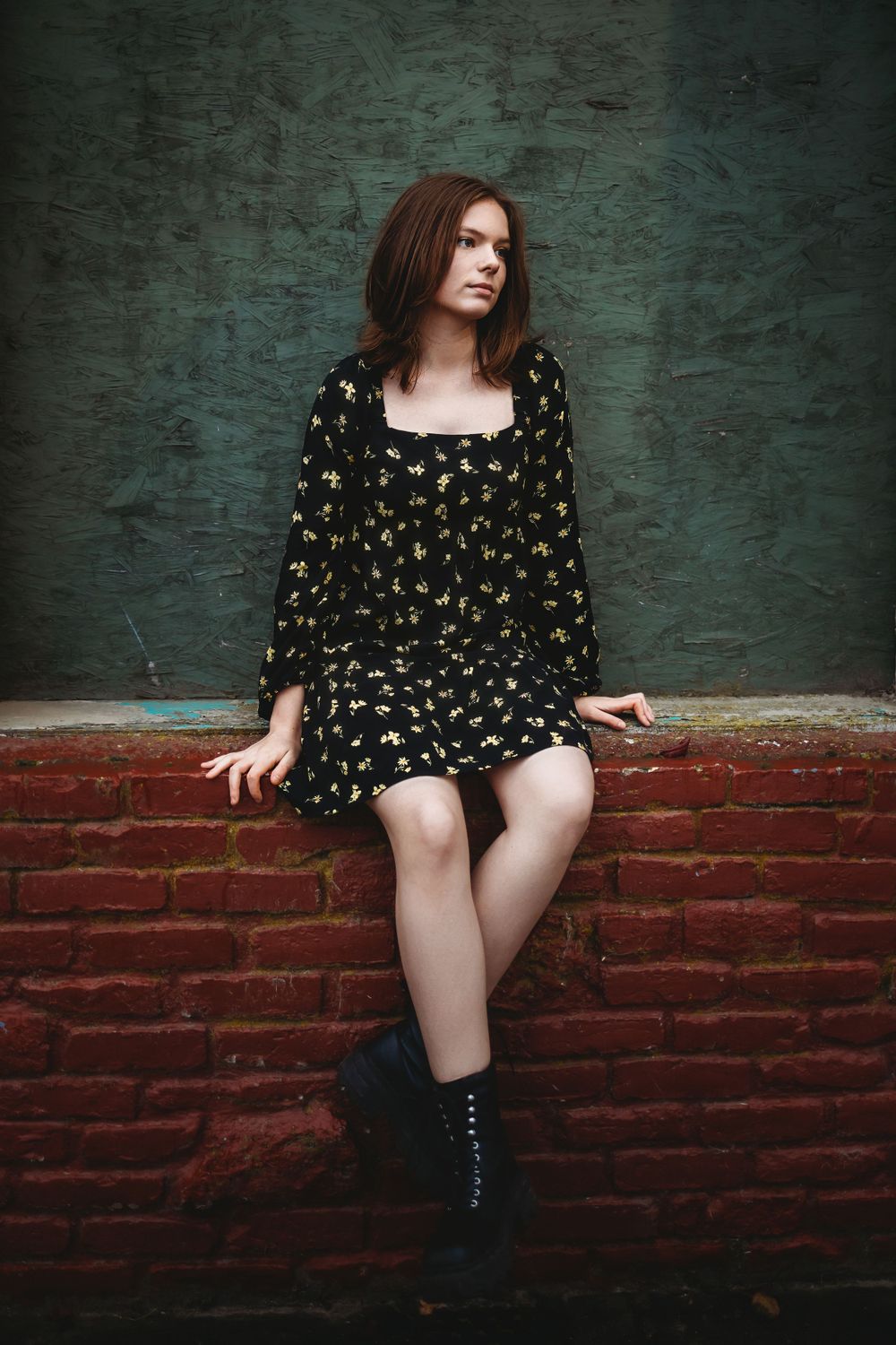 A person in a black floral dress sits on a brick ledge against a textured teal wall, wearing black boots.