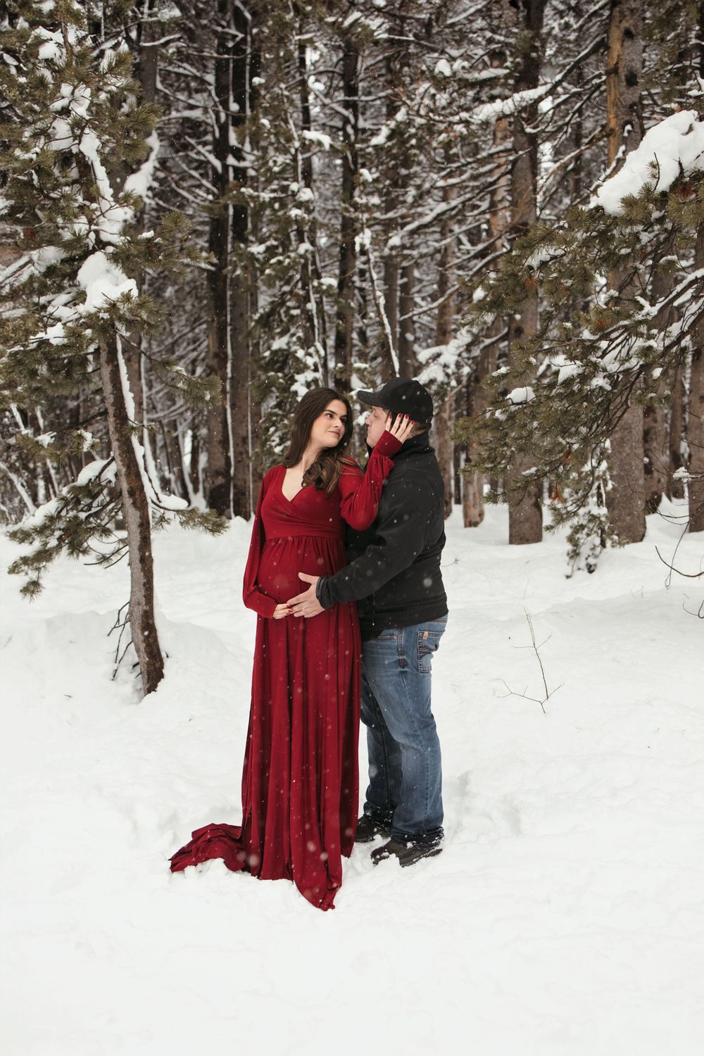 A couple embraces in a snowy forest setting with evergreen trees, the woman wearing a flowing red dress against the white snow.