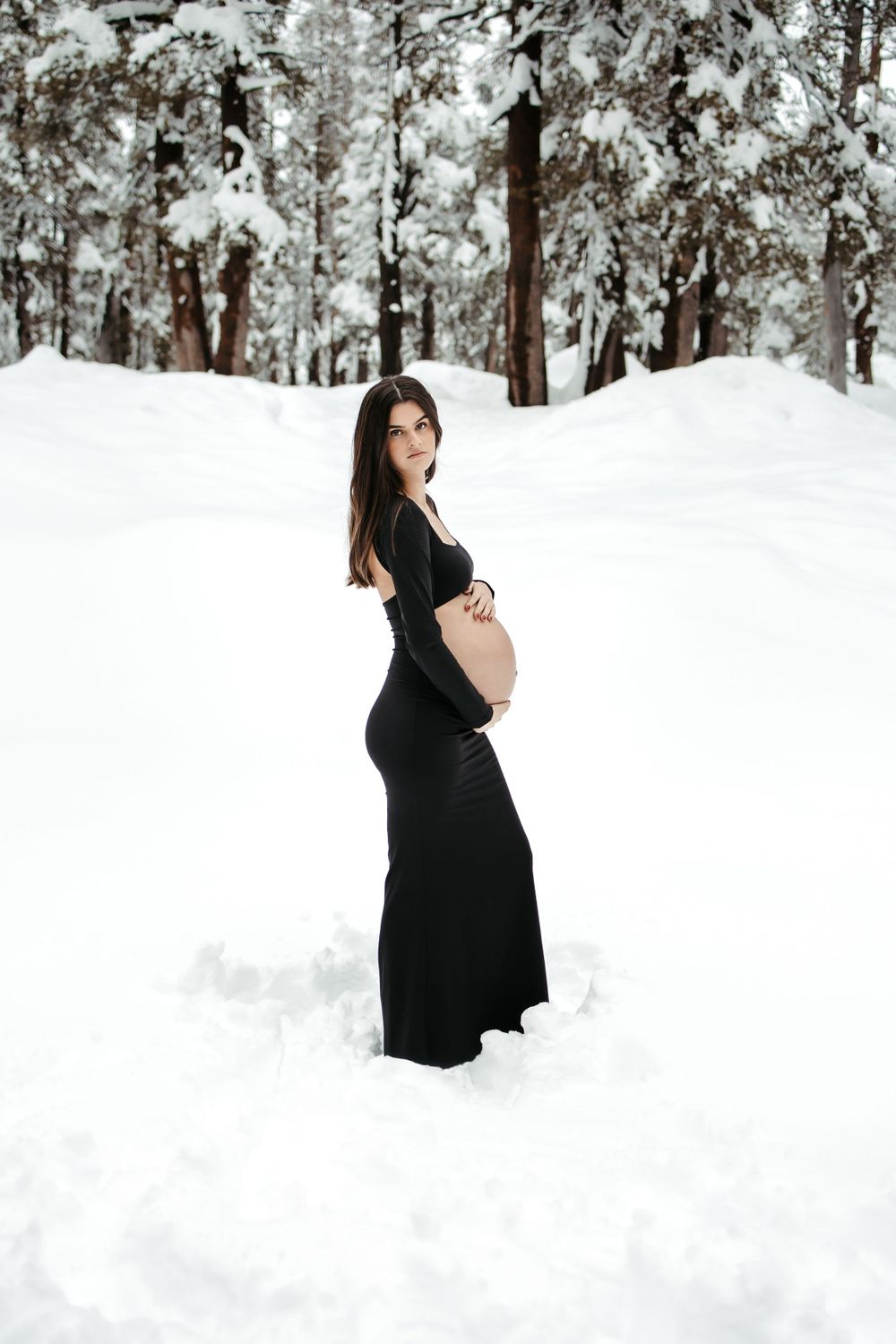 Elegant maternity photo of a figure in a long black dress standing in a snowy winter forest landscape.