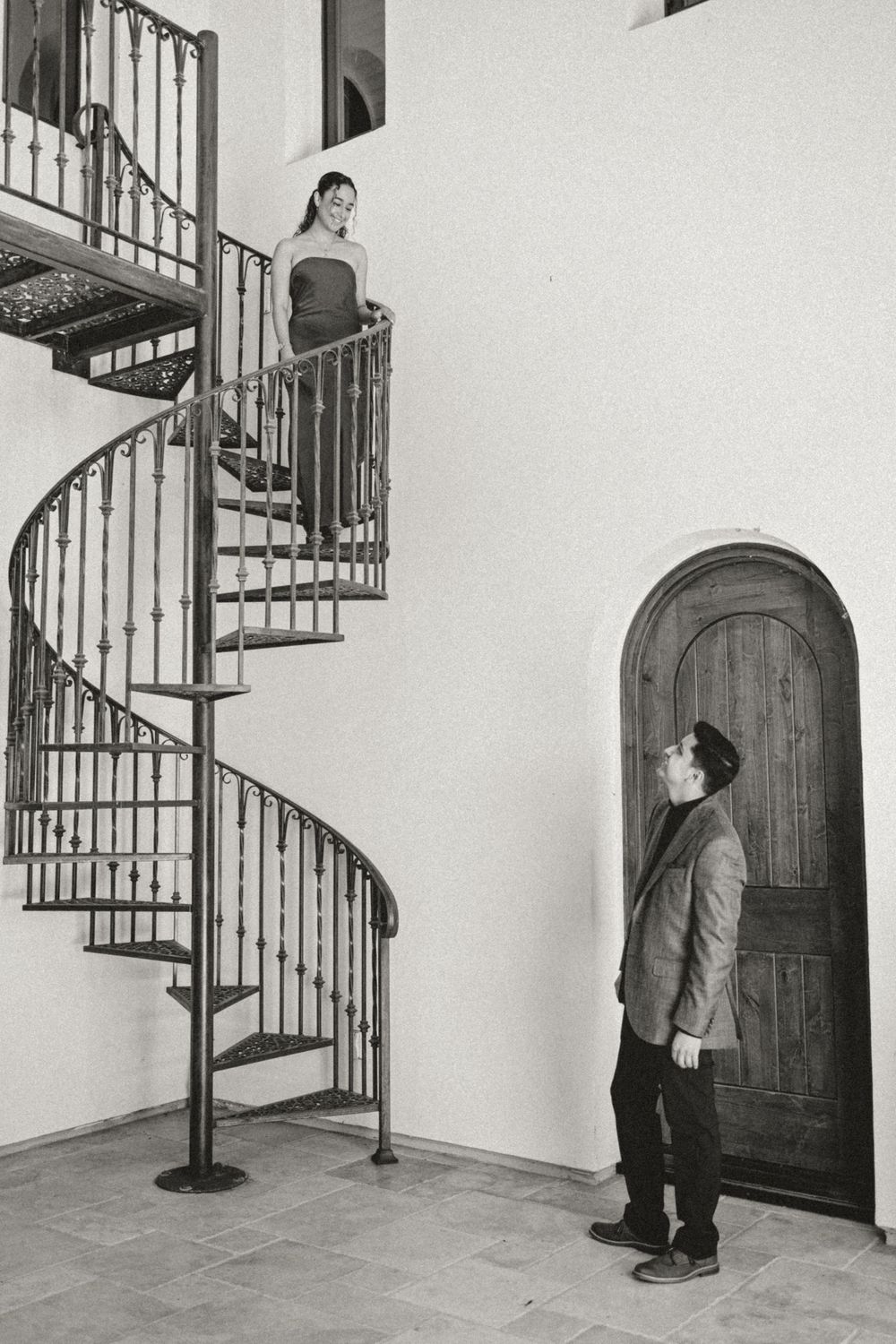 Two figures gaze at each other across a dramatic spiral staircase in a black and white architectural photograph.