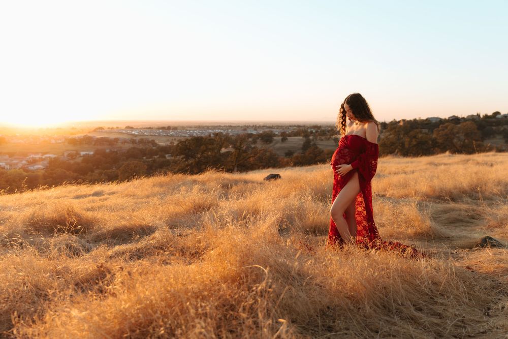 A person in a red dress stands amidst golden grass on a hilltop during sunset.