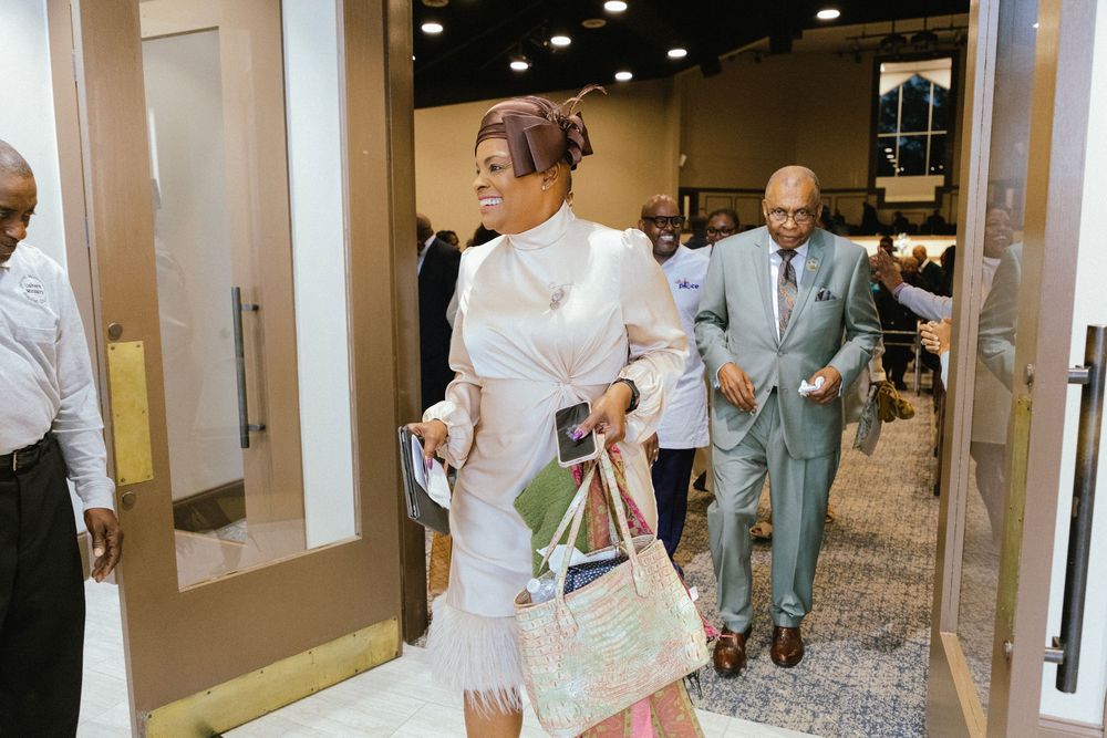 A wedding party walks through a modern event space hallway with glass walls and doors in elegant attire.