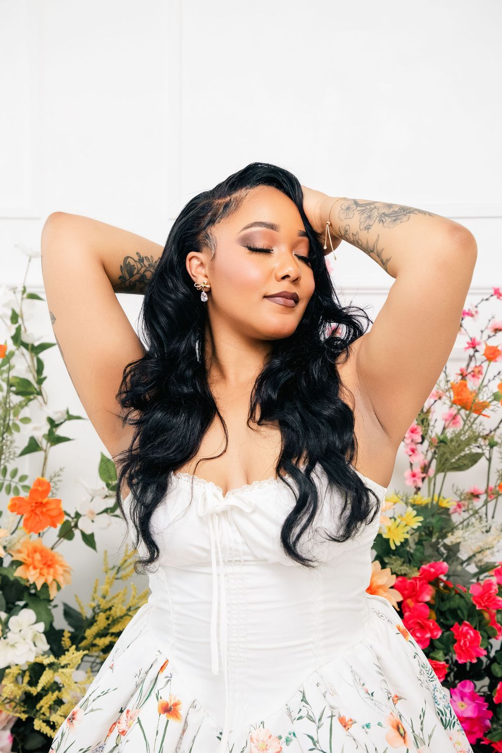 Woman in white corset dress poses with eyes closed against floral backdrop of colorful garden flowers.
