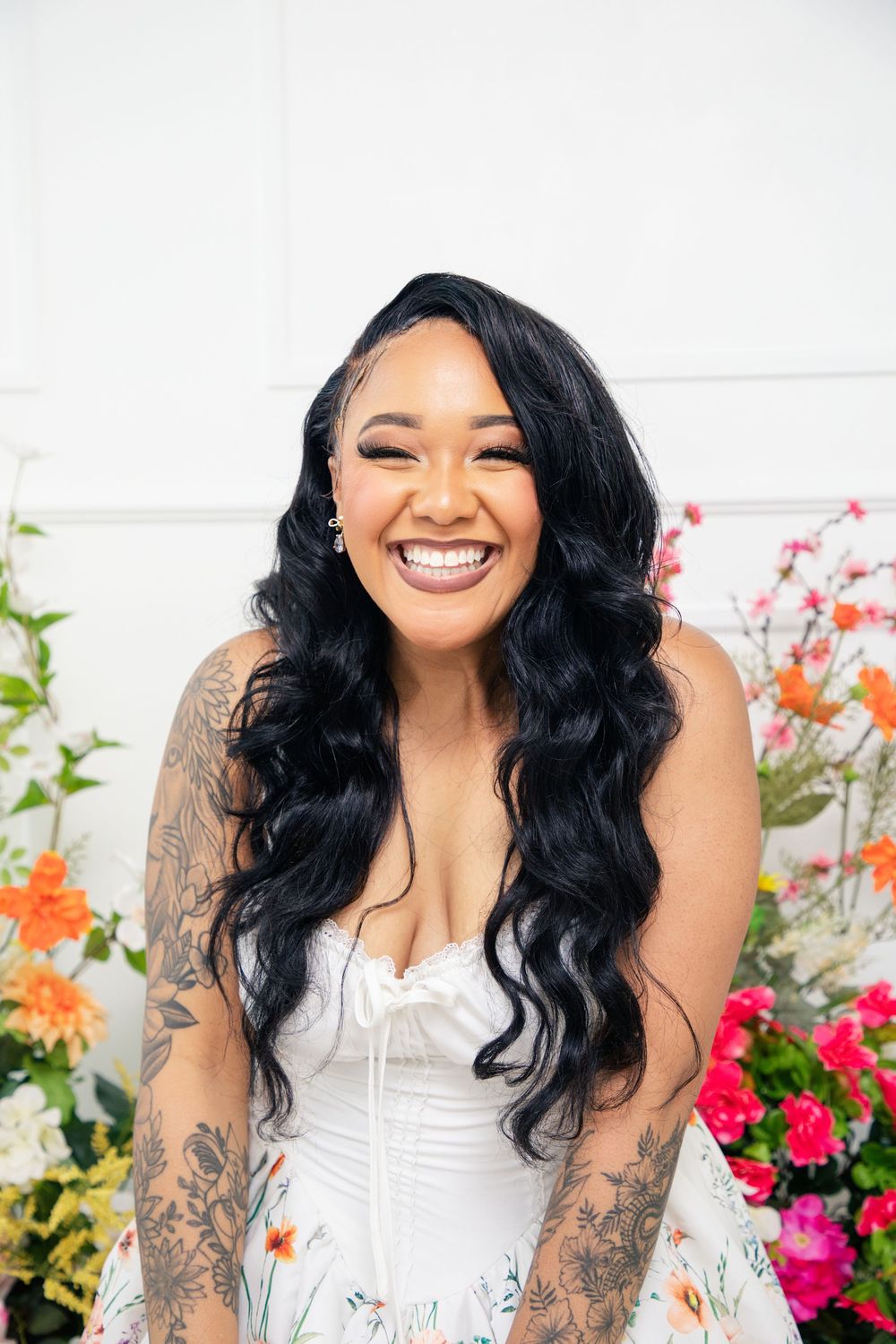 A joyful person with long wavy black hair wearing a white corset top poses against a backdrop of colorful flowers.