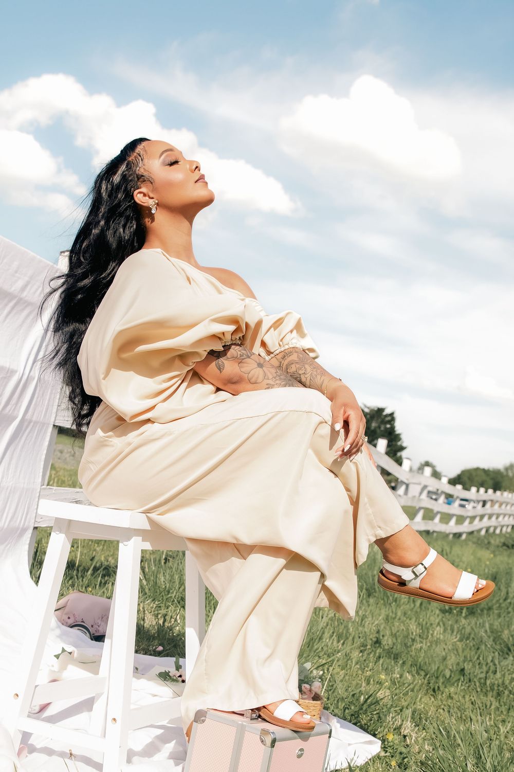 A woman in a cream-colored dress poses artistically on a white chair in a sun-lit meadow setting.