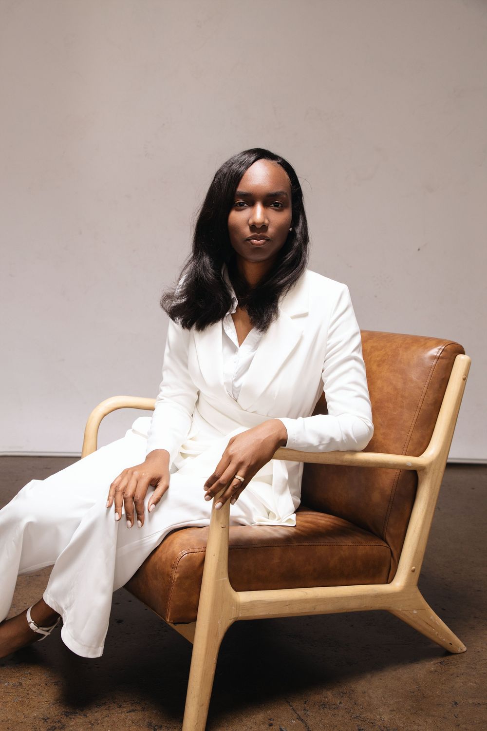 A person in a crisp white pantsuit sits elegantly in a modern brown leather and wood armchair against a neutral background.