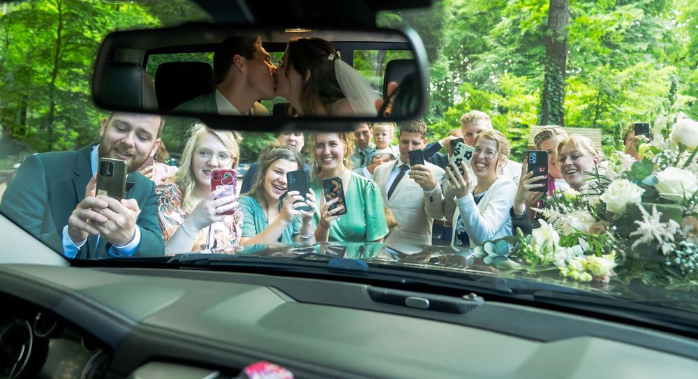 Bruidspaar lacht in de spiegel van de auto wanneer de gasten een foto maken van het paar