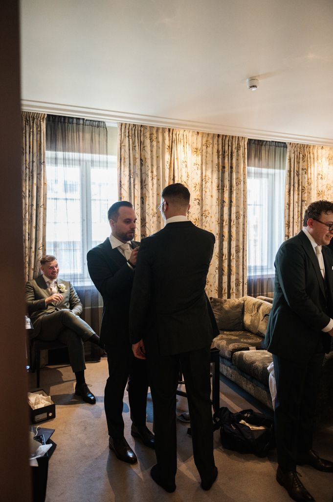 Men in formal suits gather in a hotel room with elegant curtains and furnishings before an event.