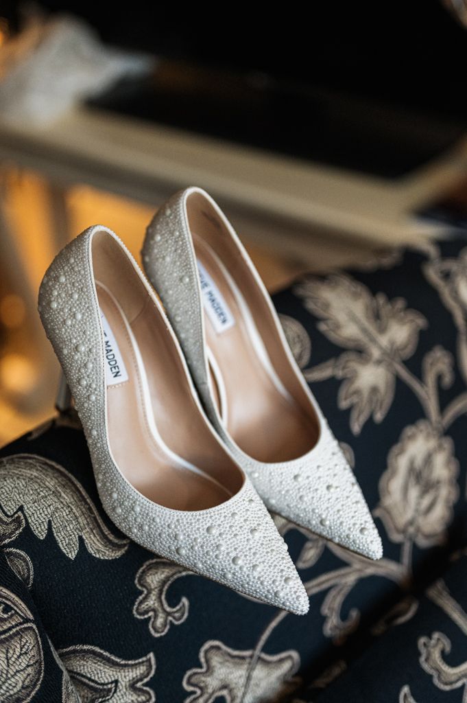 Sparkly white bridal shoes with crystal details displayed on floral upholstered furniture.