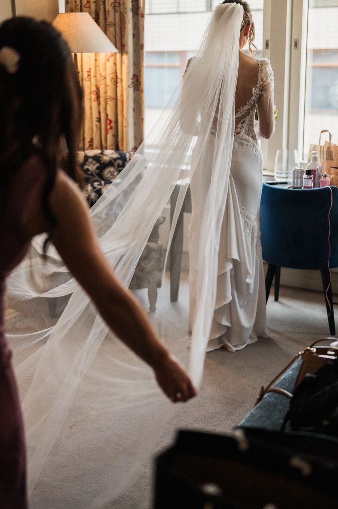 A flowing white veil trails behind an elegant lace wedding dress in a sunlit room.