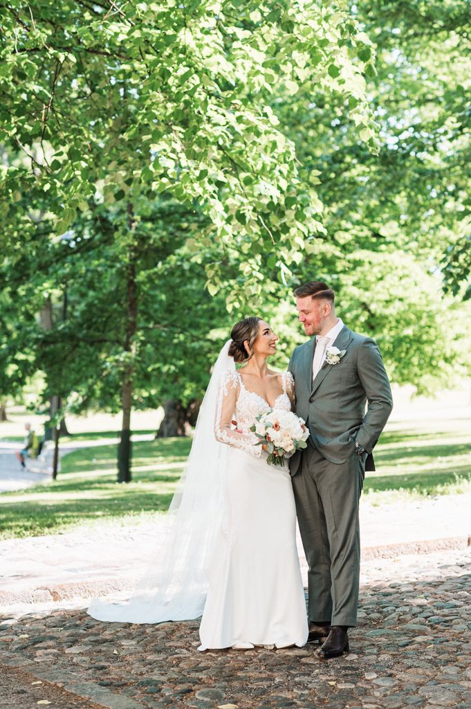 A couple in wedding attire share a joyful moment standing on a cobblestone path surrounded by lush green trees.