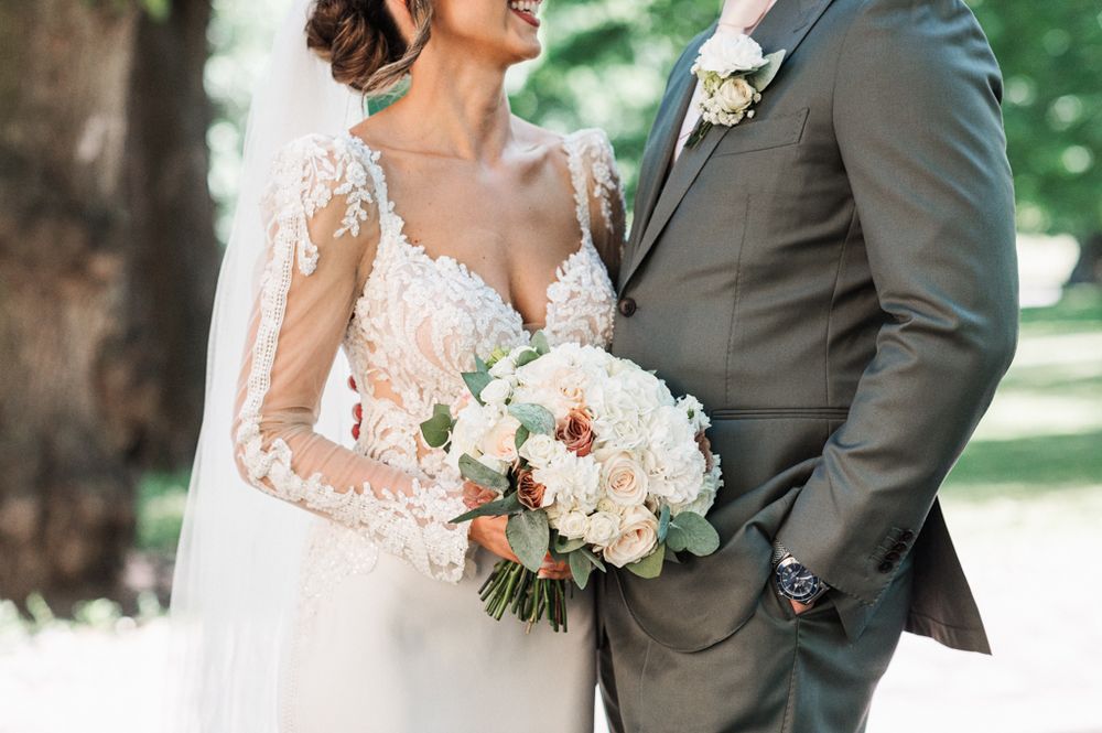 A wedding couple in elegant attire standing close together, with the bride holding a lush white and blush floral bouquet.