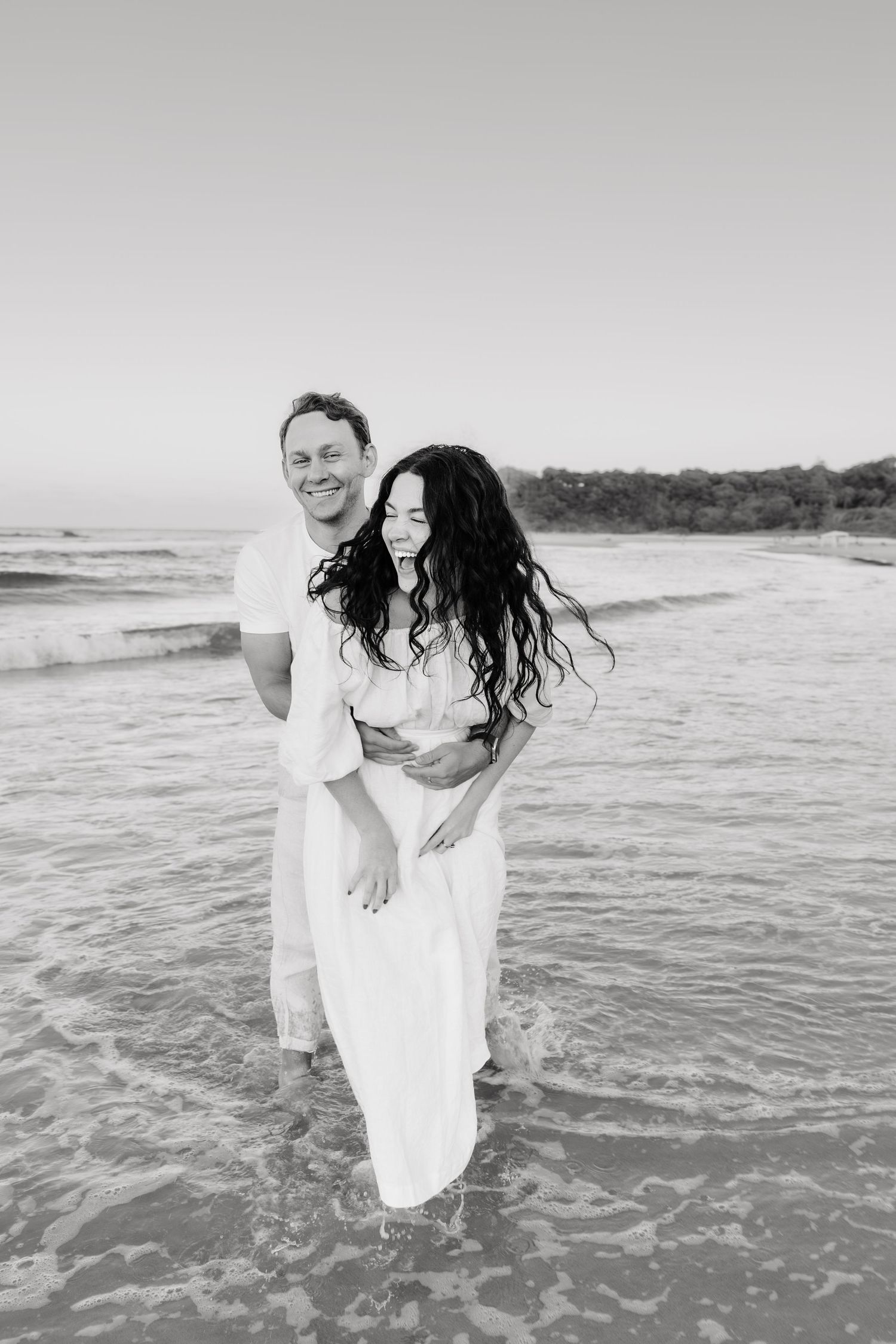 A couple shares a joyful moment together while standing in shallow ocean waves on a beach in black and white.