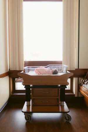 A row of four hospital cots with wooden rails in a bright room with cream curtains and natural lighting.