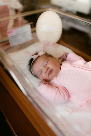 A newborn baby sleeping peacefully in a hospital bassinet wearing a pink outfit with a white pom-pom decoration above.
