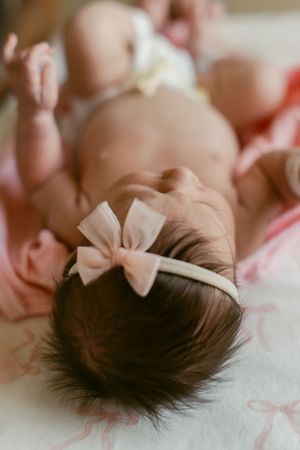 A sleeping baby wearing a pale pink bow headband lies on soft fabric, delicate lighting highlighting their dark hair.