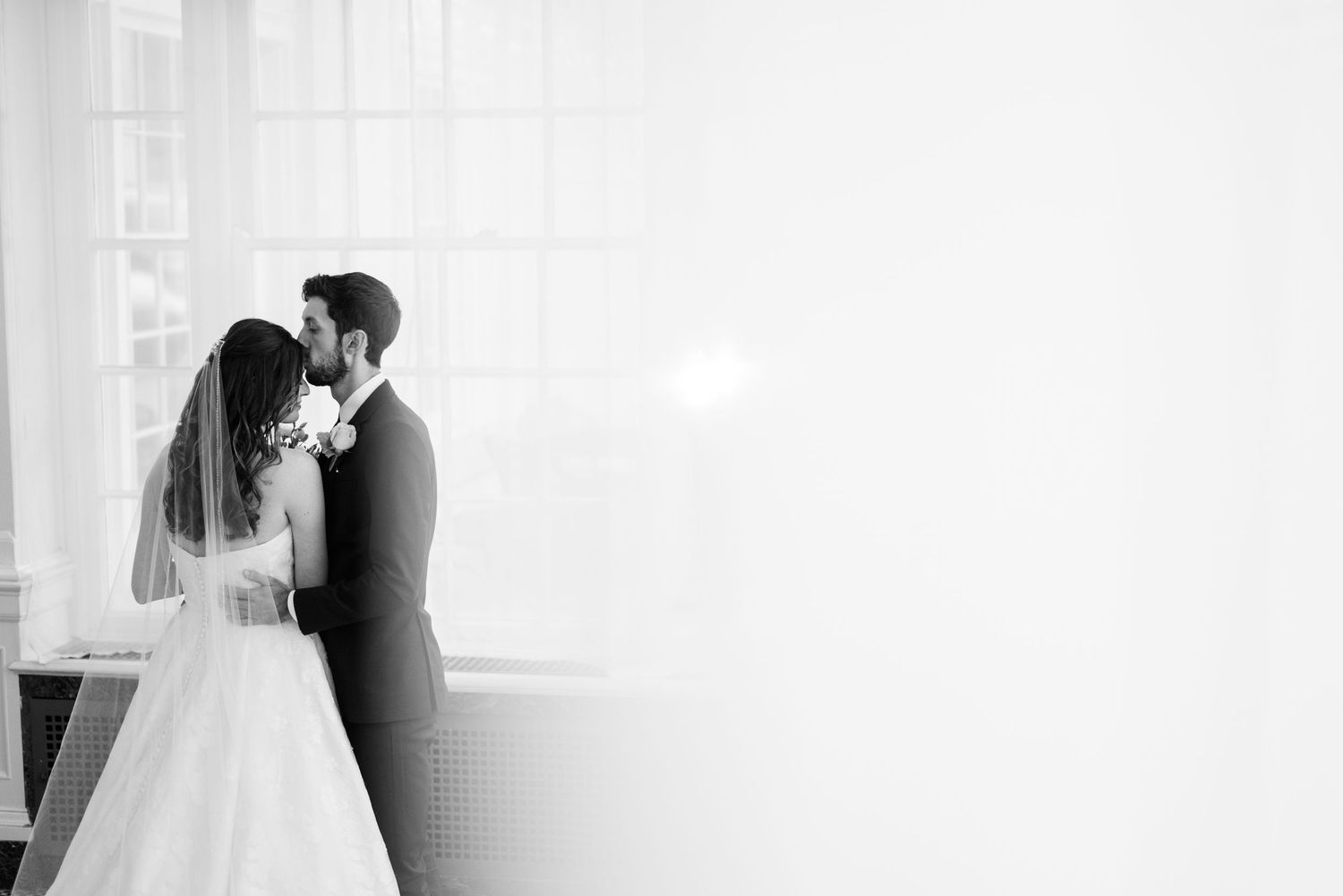A romantic black and white wedding photograph of a couple embracing by a window with soft natural light