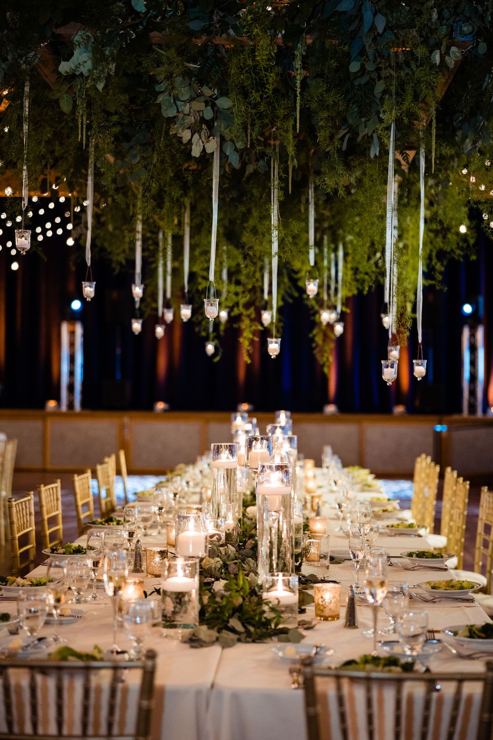 Elegant wedding reception table with candles, greenery centerpieces, and hanging lanterns beneath a lush floral ceiling.