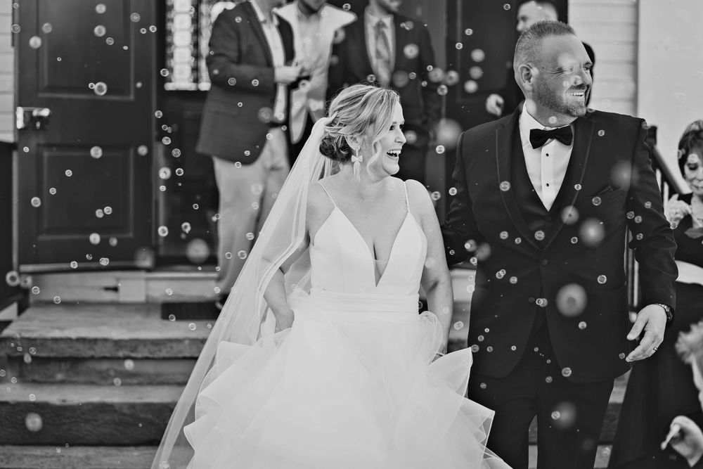 Black and white photo of a bride and her father walking out of a church surrounded by bubbles on her wedding day.