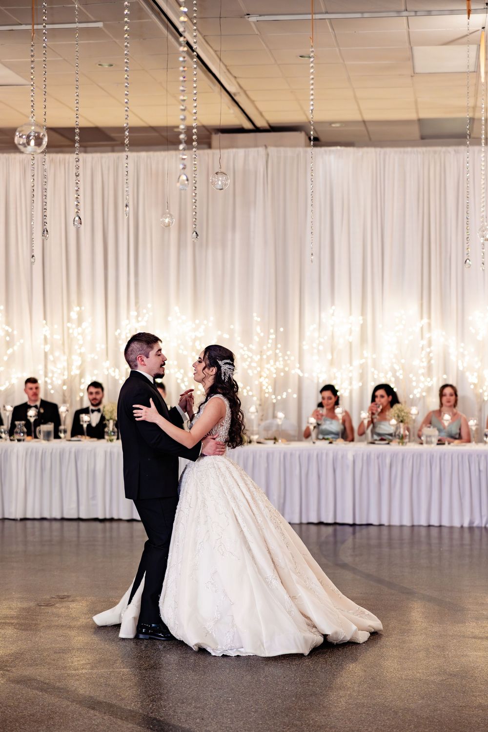 Bride and groom share their first dance at elegant wedding reception with crystal decor and glowing white backdrop.
