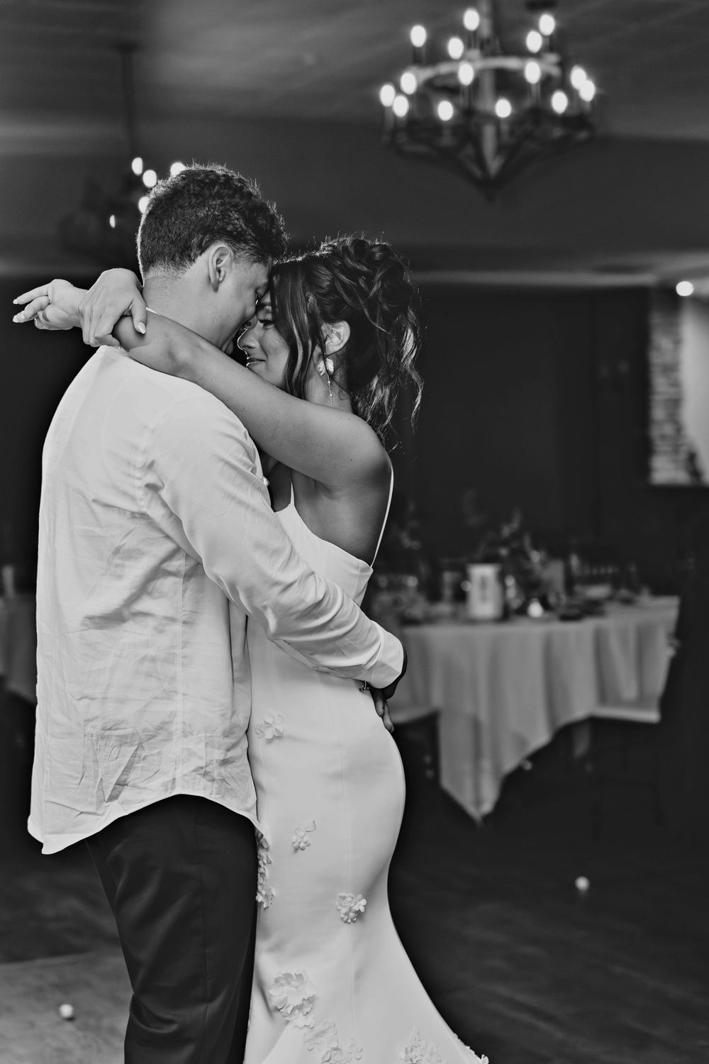 Black and white photo of bride and groom sharing first dance, embracing tenderly under chandelier at wedding reception.