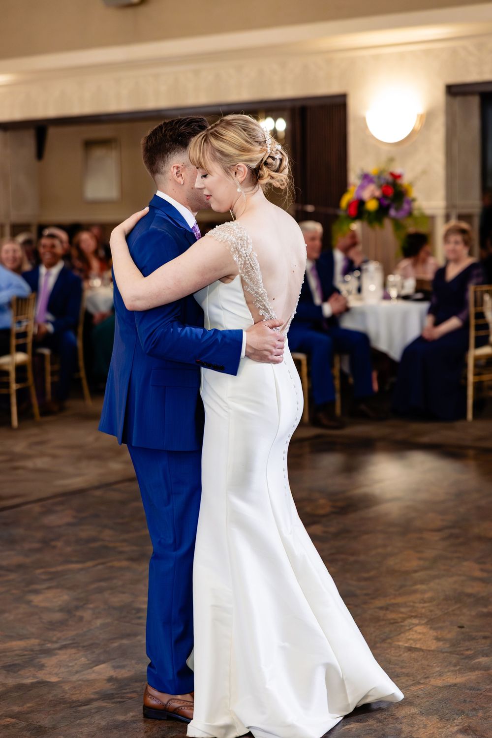 Bride and groom share their first dance in a wedding reception hall, guests seated at decorated tables in the background.