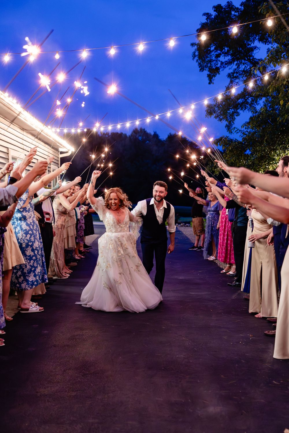 Bride and groom exit wedding reception through sparkler tunnel with string lights overhead at twilight.