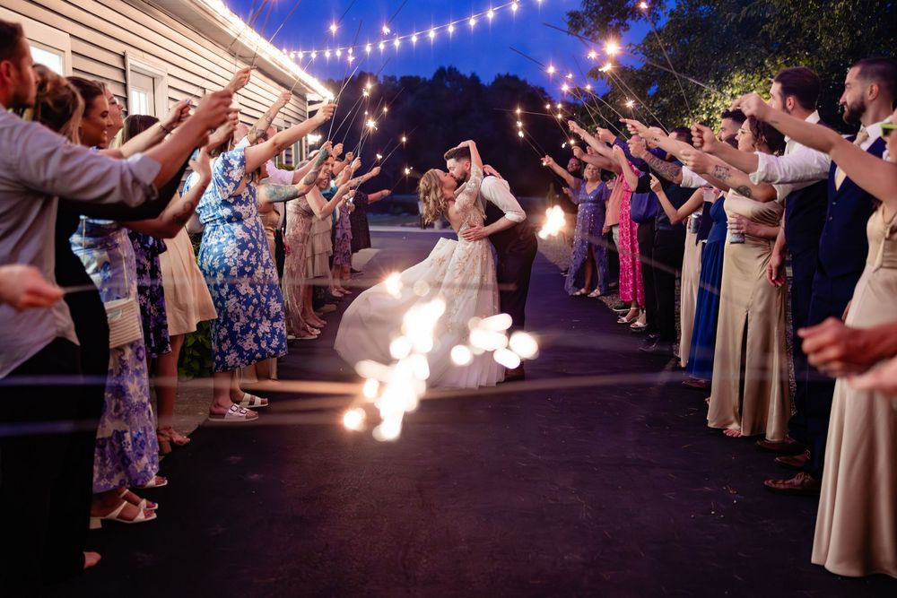 Bride and groom exit through sparkler tunnel with wedding guests at night under string lights.