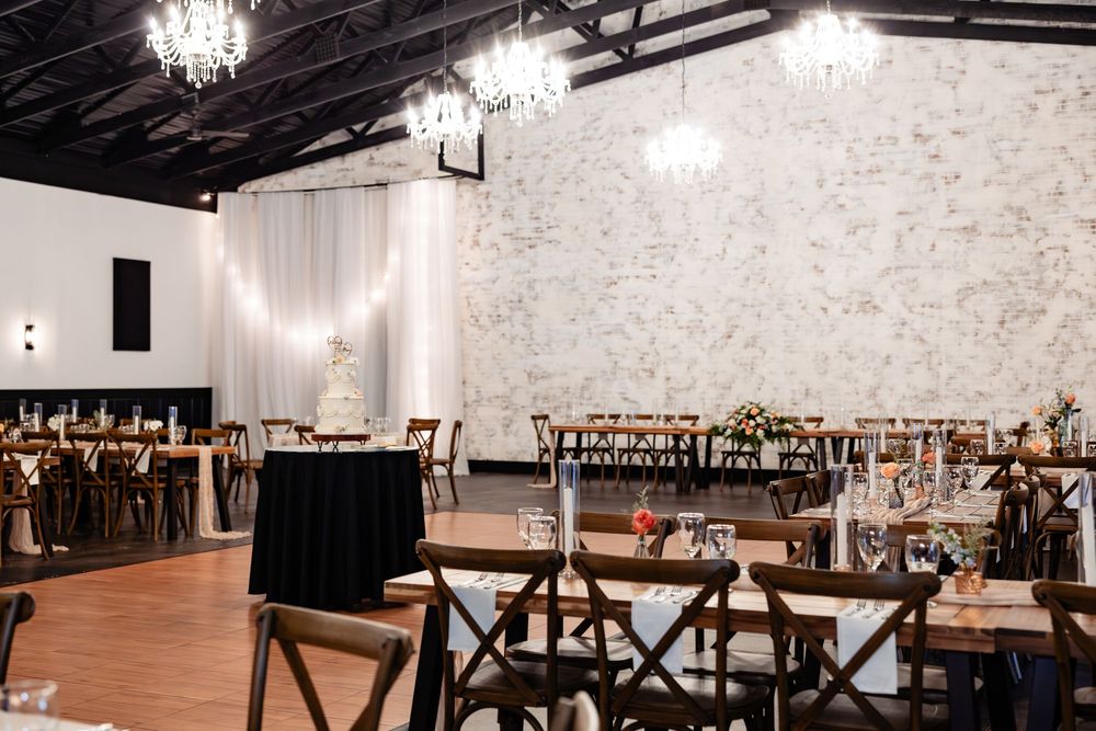 Elegant wedding reception venue with chandeliers, rustic wooden chairs, draped white fabric, and long banquet tables on hardwood floors.
