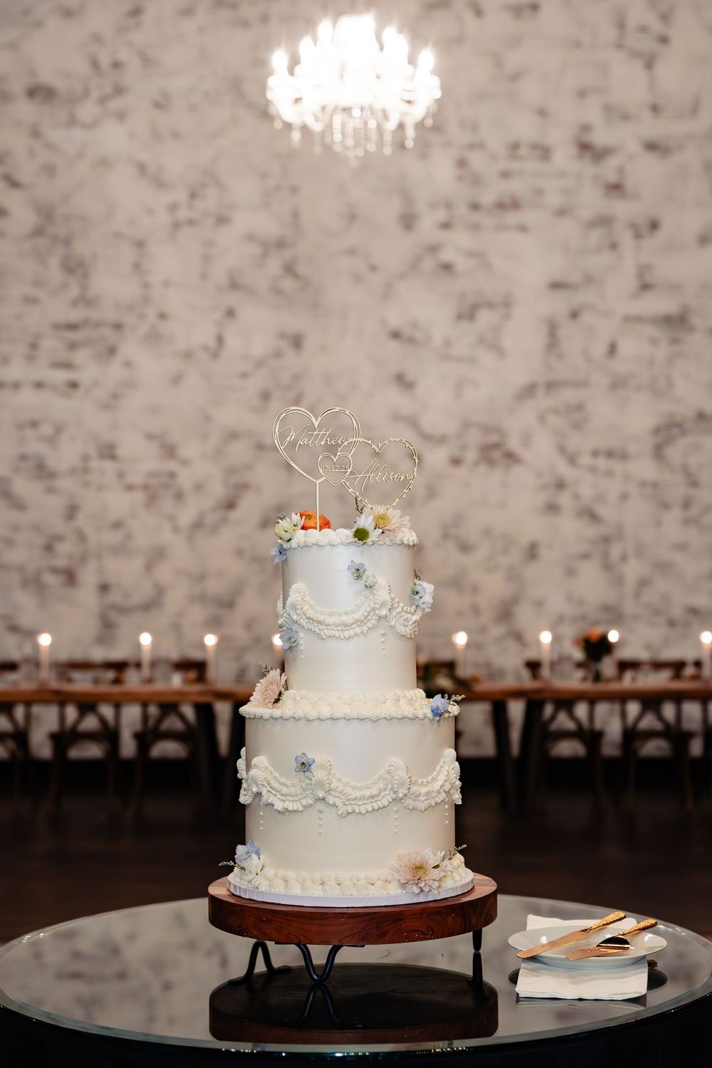 Elegant three-tier white wedding cake with floral accents and gold topper on a wooden stand in a candlelit reception hall.