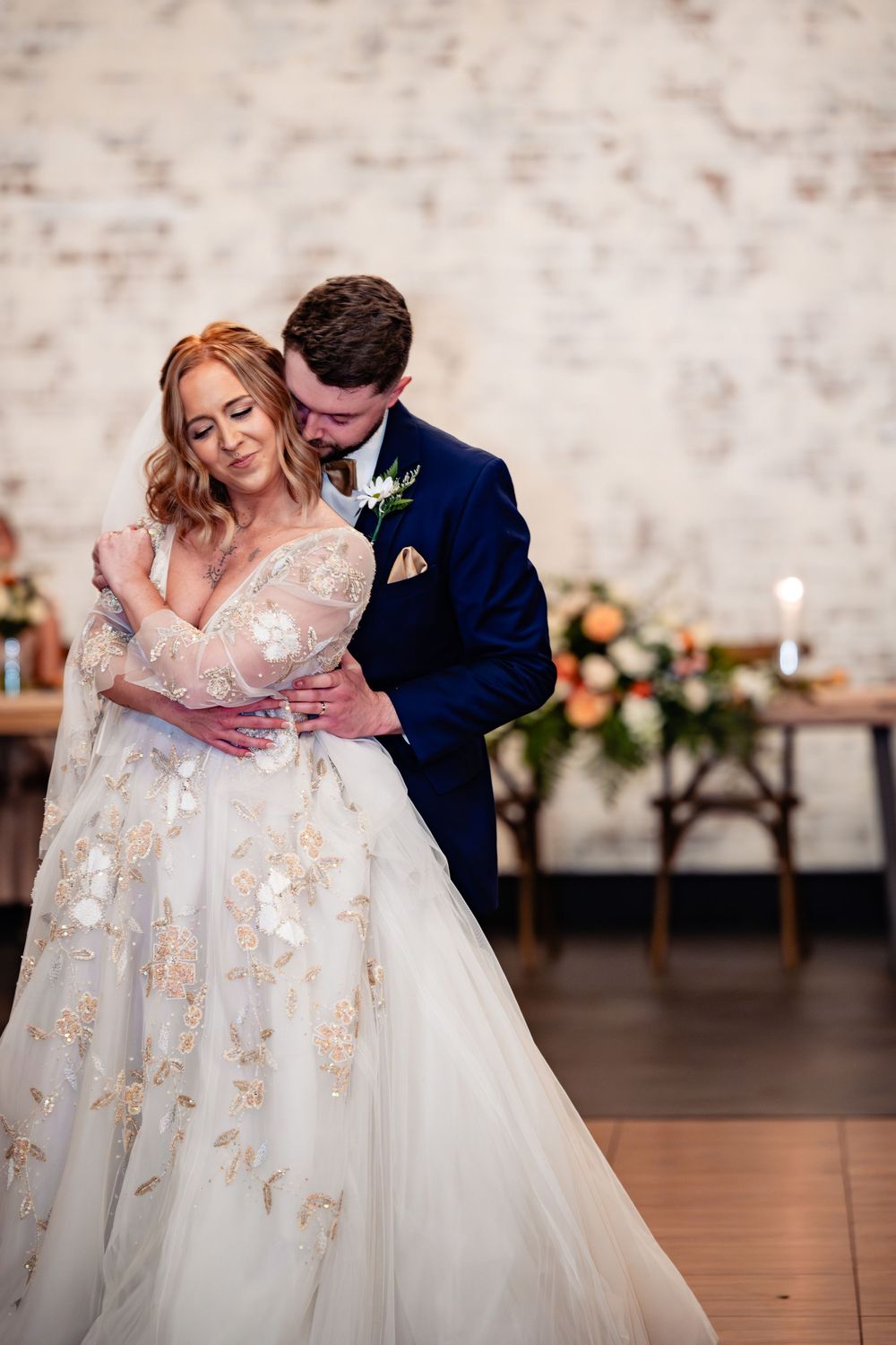 Bride in floral gown embraced by groom in navy suit during first dance at elegant wedding reception.