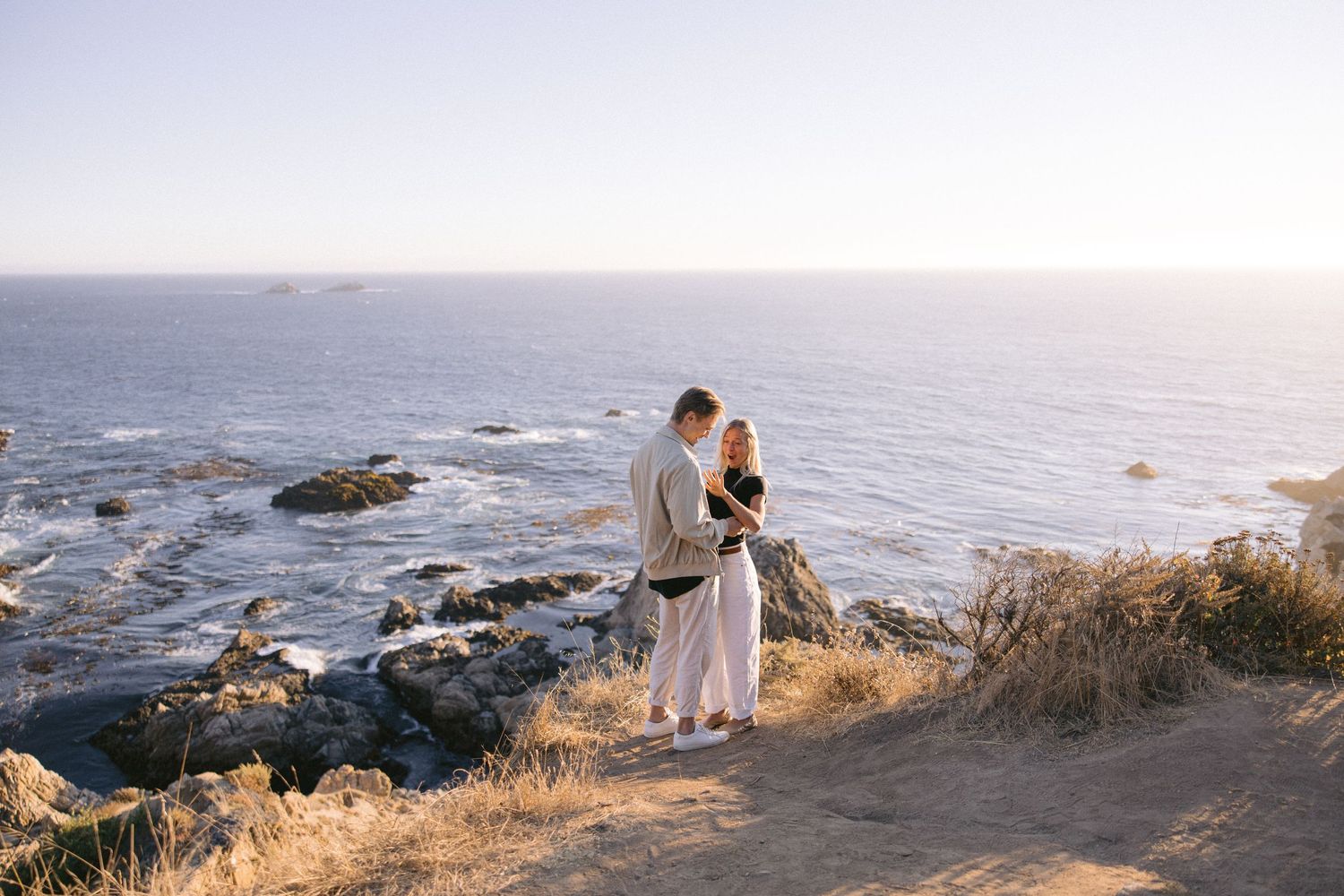 Couple stands on rocky cliff overlooking ocean during sunset along the California coastline.