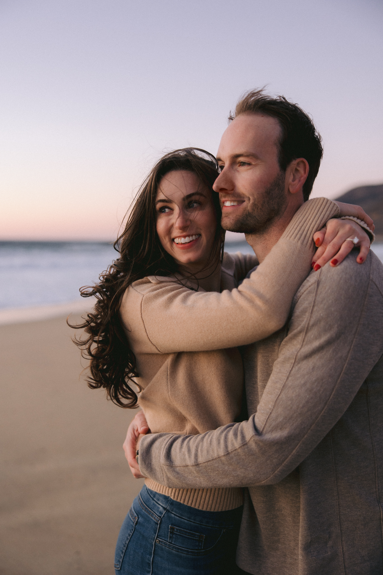 A happy couple embraces on the beach at sunset wearing neutral colored sweaters.