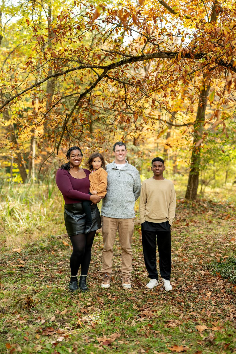 Blended family of four posing together under vibrant red and gold autumn tree branches.