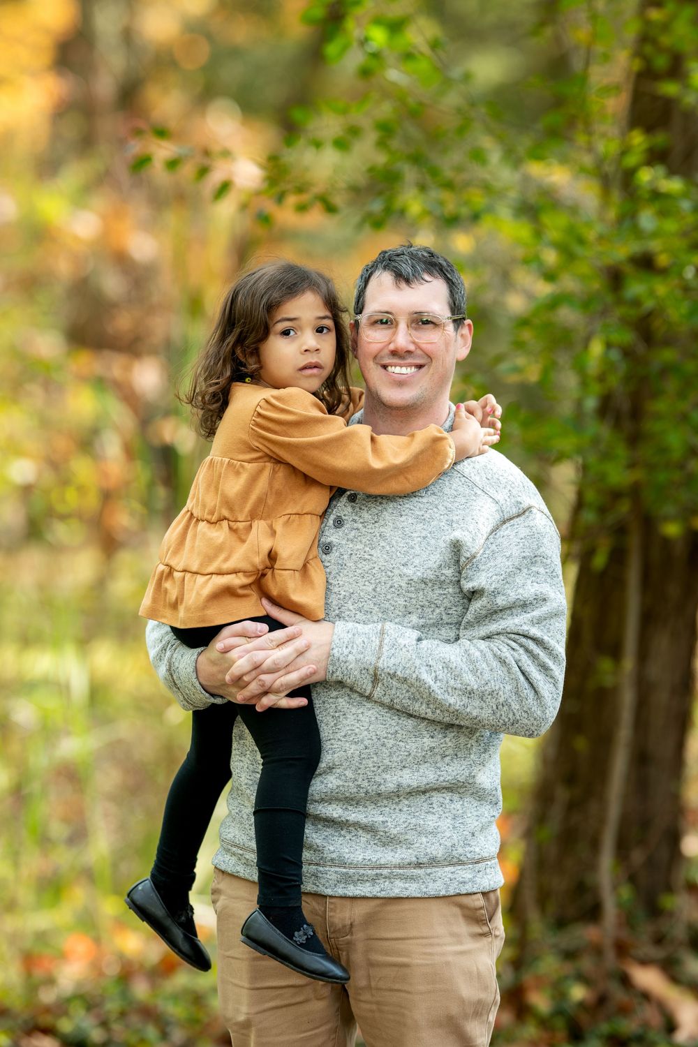 Smiling father holding his young daughter in an orange dress in a fall foliage park.