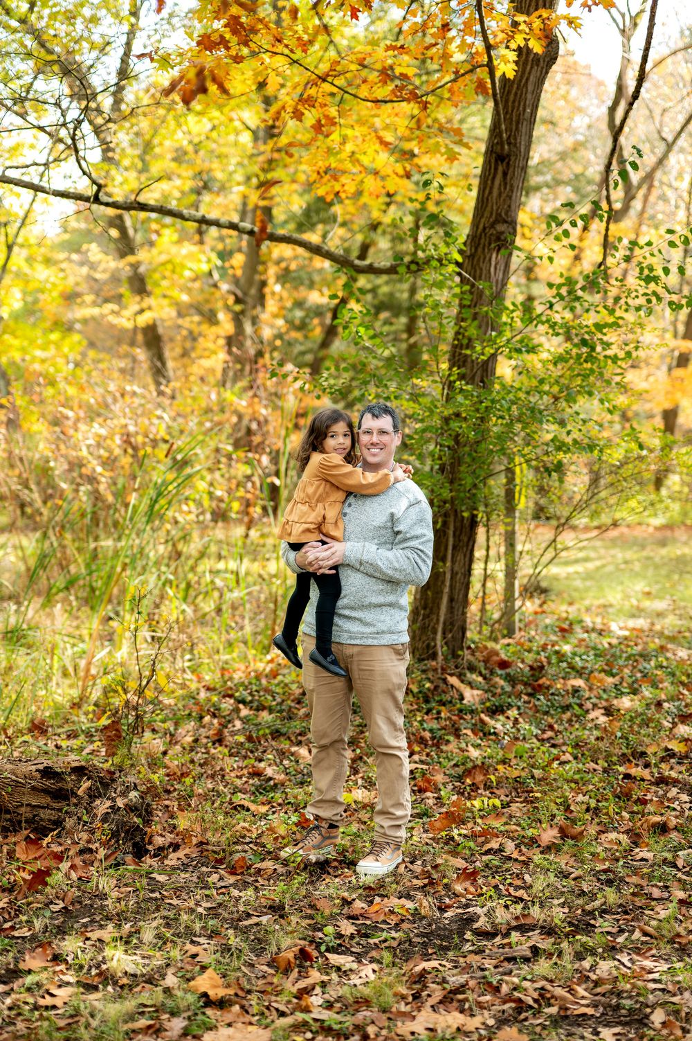 Father holding young daughter in orange dress while smiling in a vibrant autumn forest.