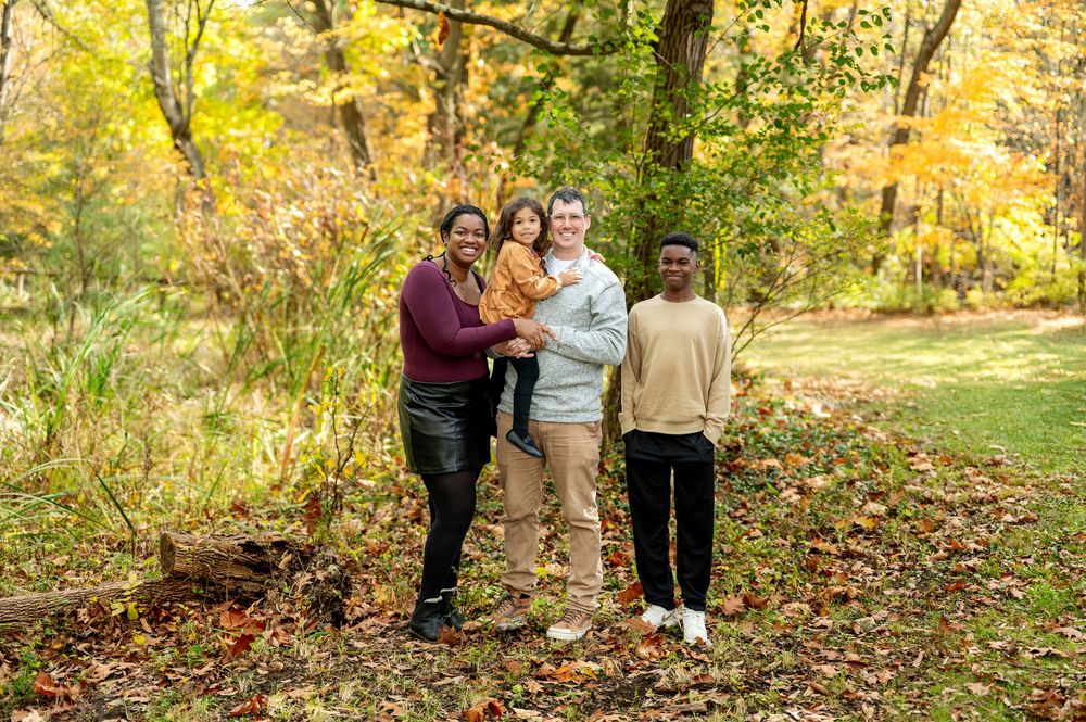 Smiling blended family of four posing together on a leaf-covered path in autumn woods.